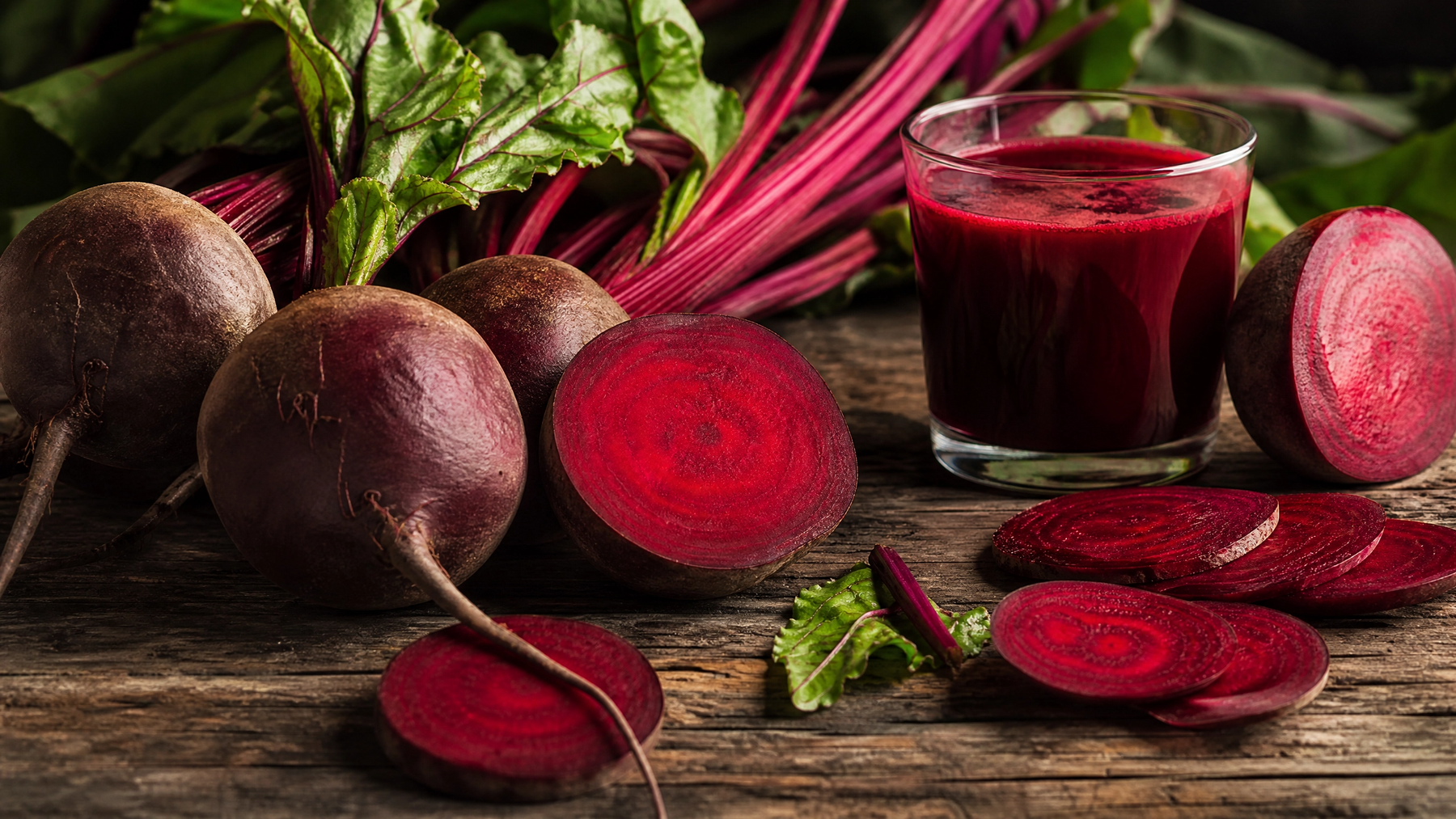 Fresh whole and halved beetroots with leafy tops, sliced beetroot rounds and a glass of deep red beetroot juice on wood