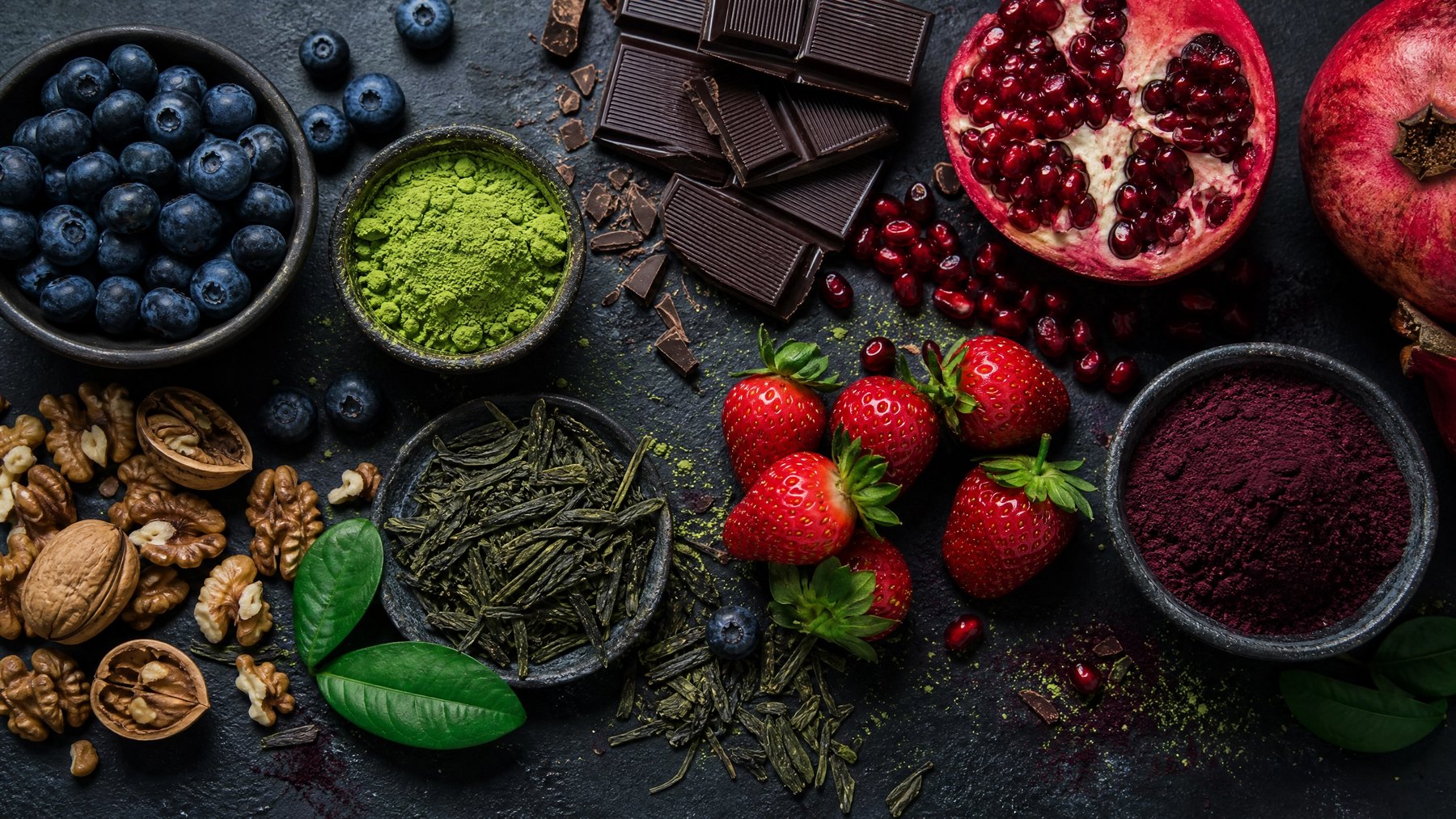 Antioxidant-rich foods including blueberries, pomegranate, dark chocolate, matcha powder and strawberries on dark slate